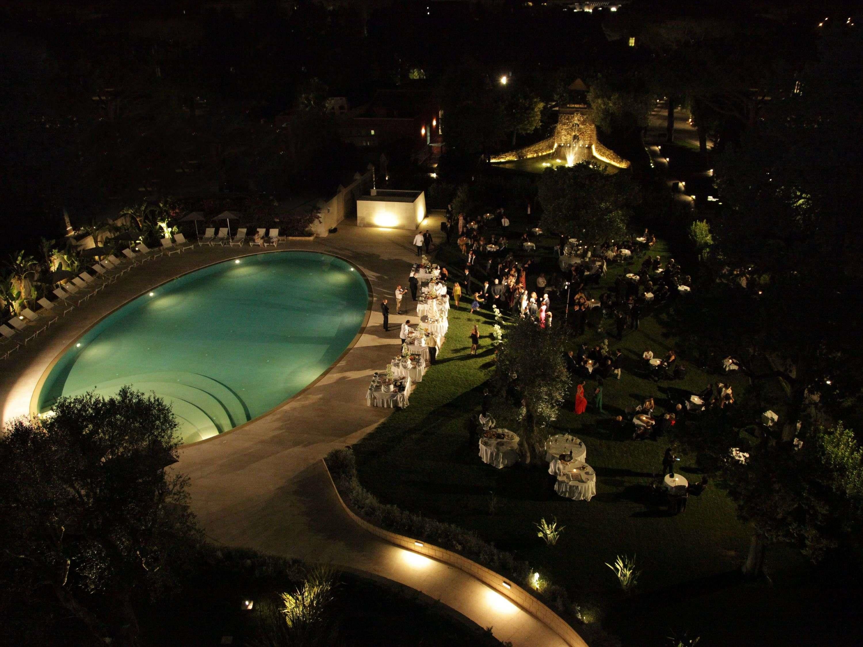 Vista aérea de un complejo turístico con una gran piscina, zonas de comedor al aire libre y un camino bien iluminado.