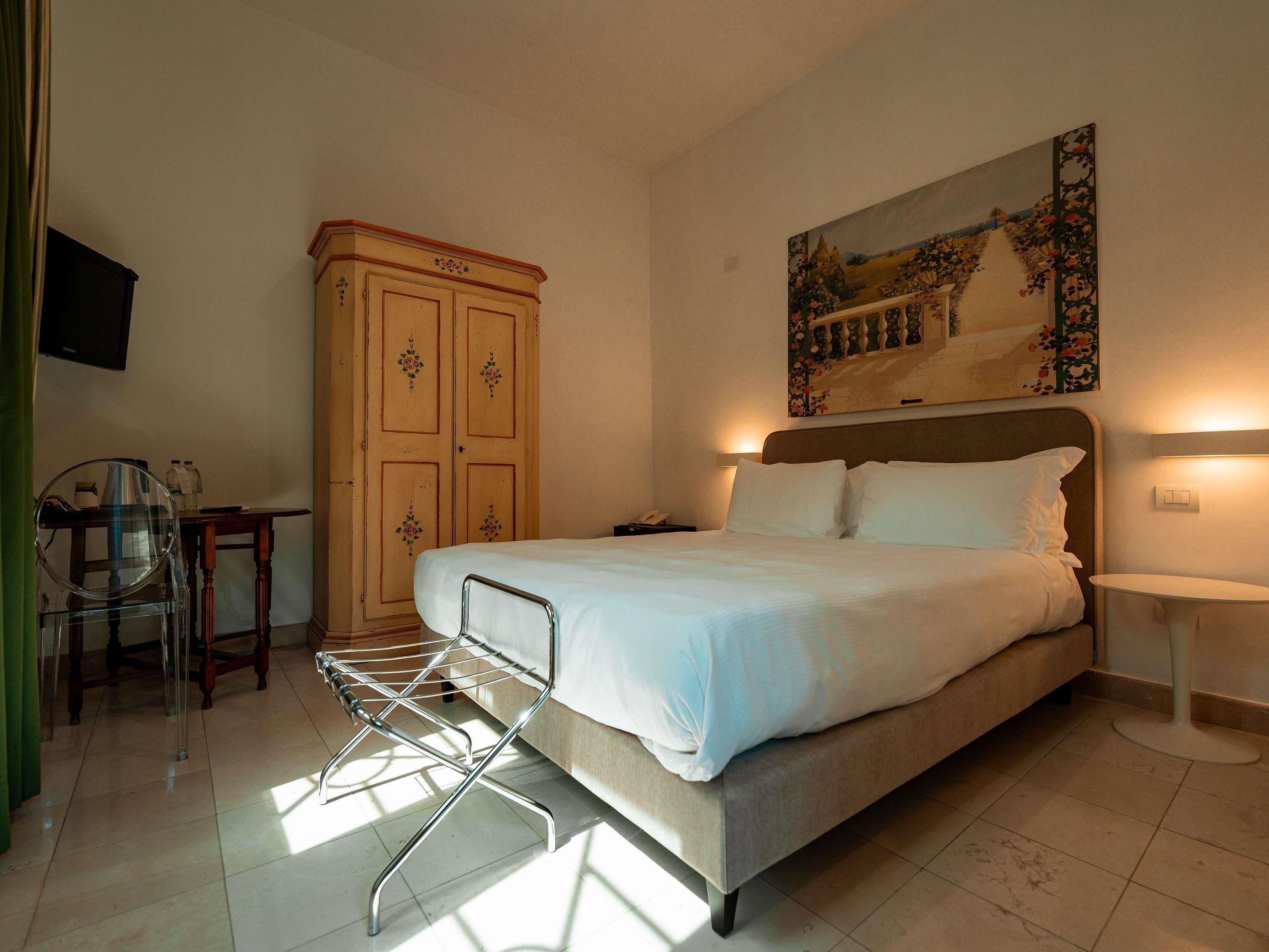 A hotel room with a bed, a wardrobe, a desk with a chair, and a painting on the wall.