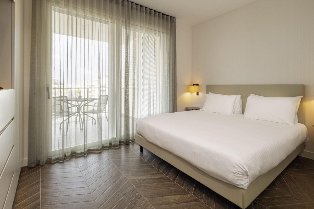 A hotel room with a large bed, a small table, and a view through sheer curtains.