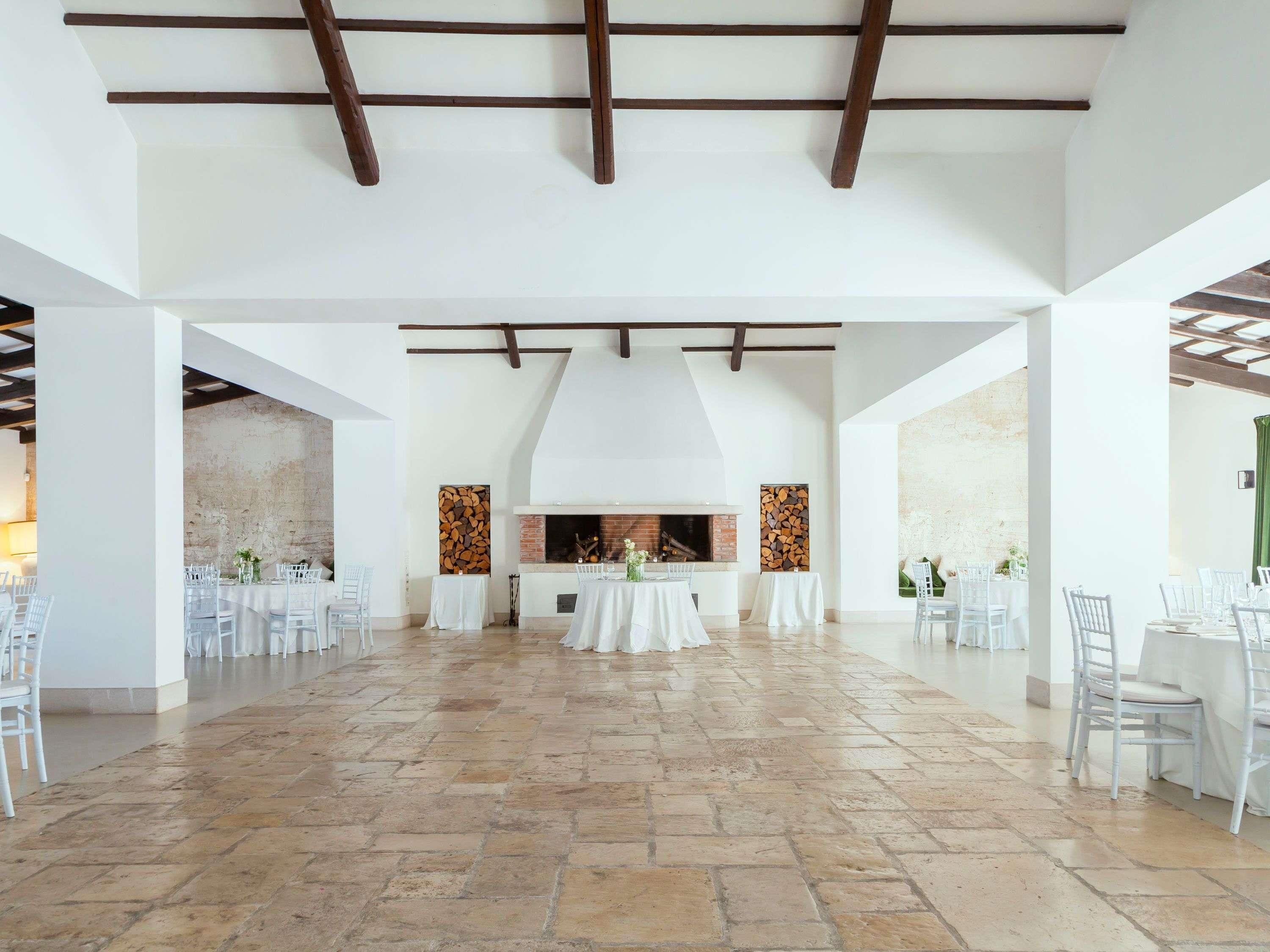 A spacious hall with a high ceiling, exposed beams, and a central fireplace. Tables are set with white tablecloths and chairs.