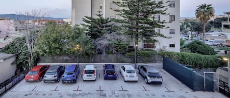 Een parkeerplaats met verschillende auto's, een hoog gebouw en een grote boom op de voorgrond.