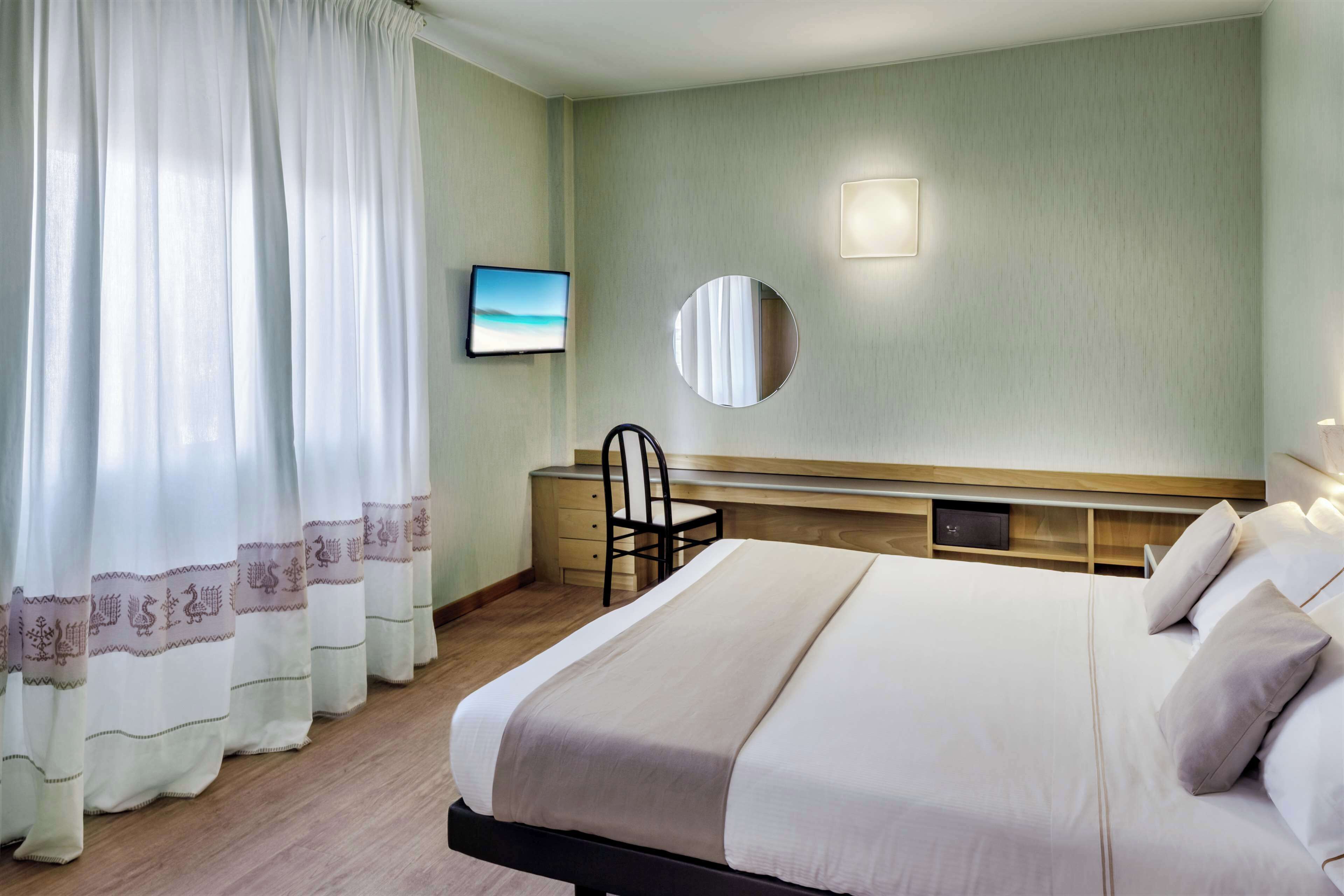 A hotel room with a bed, a desk, a chair, a TV, and a wall-mounted mirror.
