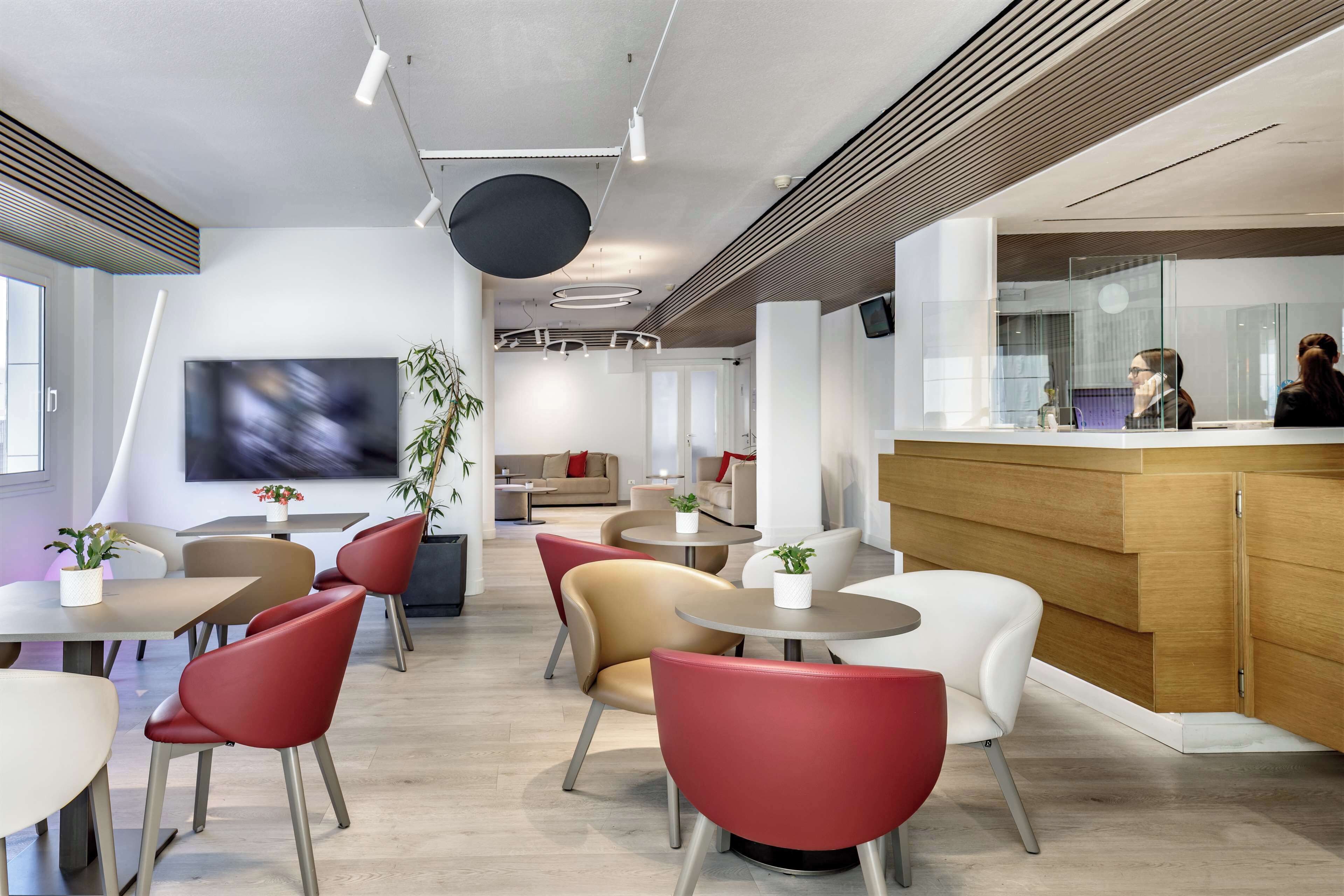 Eine moderne Hotelhalle mit runden Tischen, roten und beigen Sesseln, einem Empfangsbereich und einem großen Flachbildfernseher.