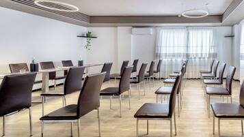 A conference room with rows of chairs and tables, a ceiling with circular light fixtures, and a large window with curtains.