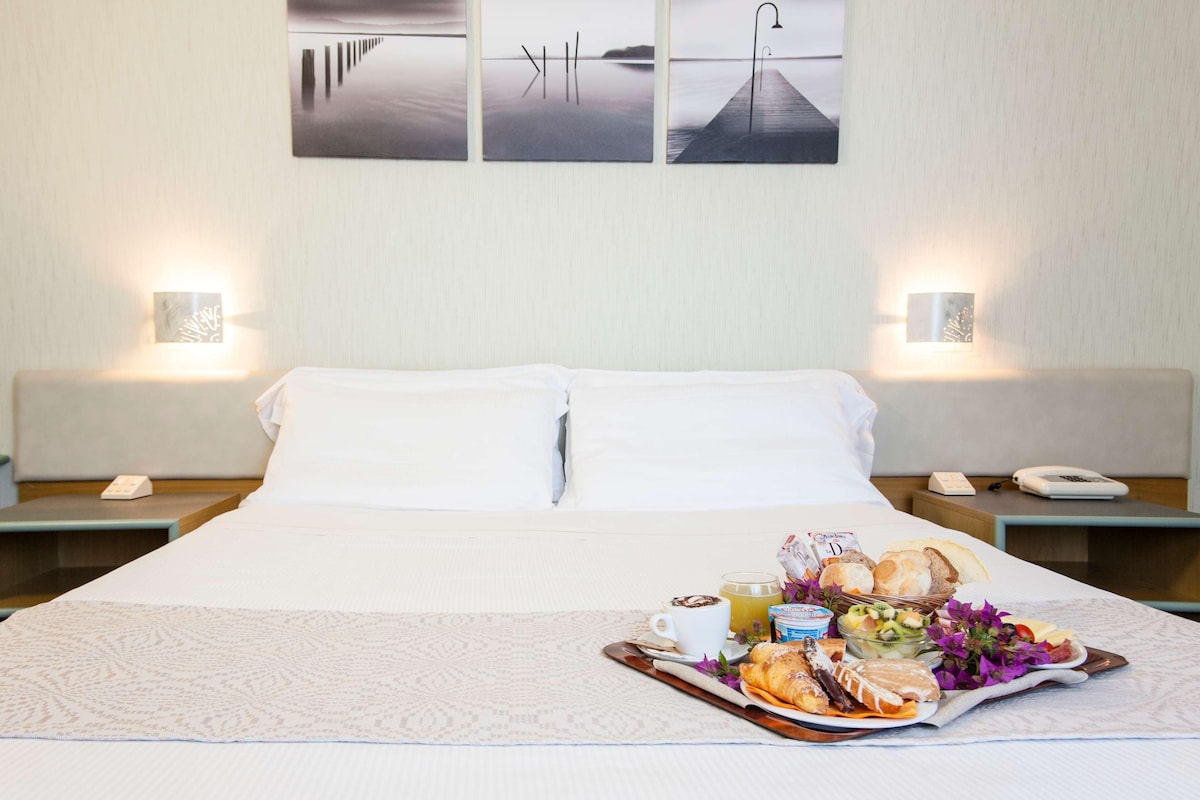 Habitación de hotel con una cama bien hecha, una bandeja de desayuno con croissants, fruta y café, y dos lámparas fijadas a la pared.