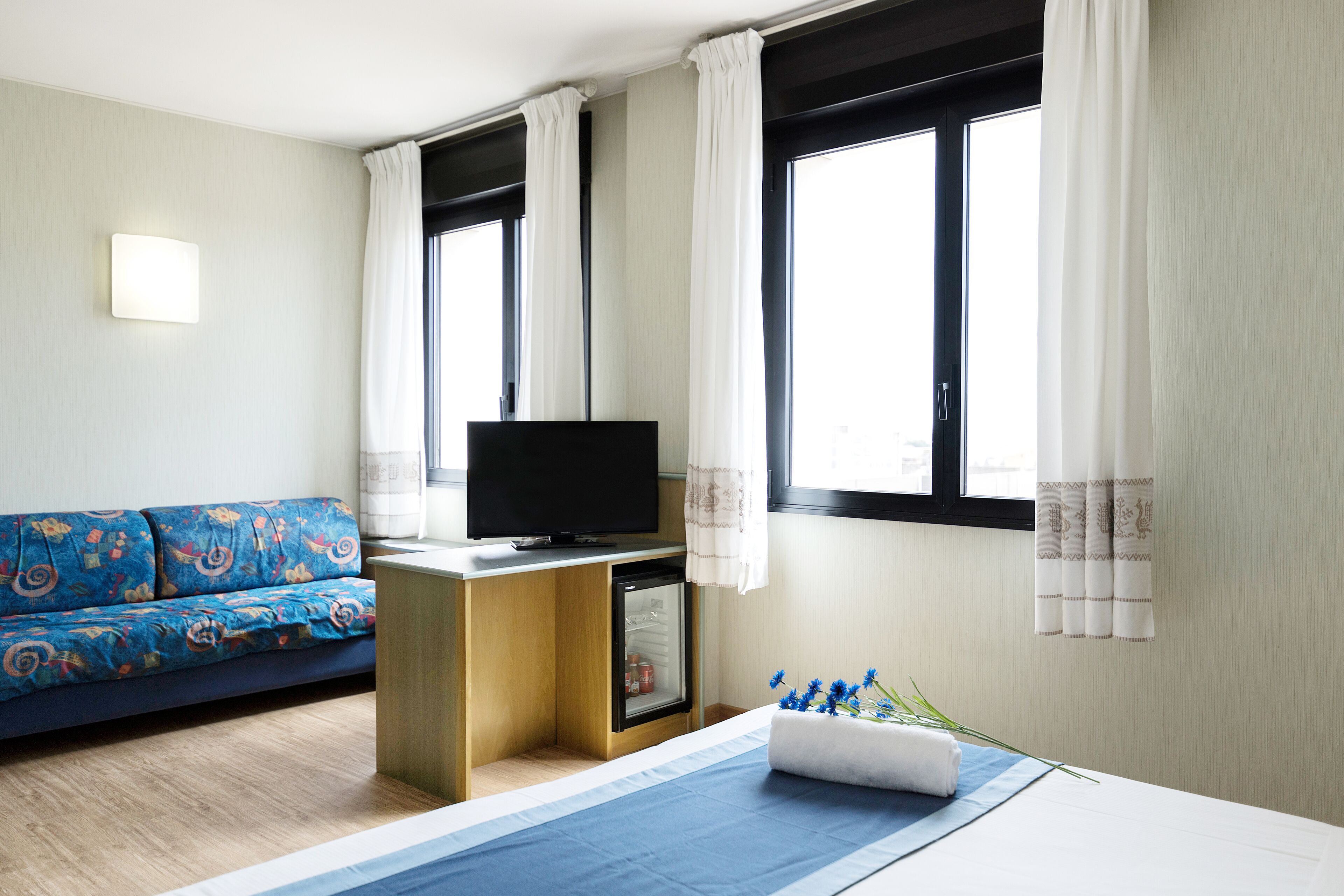 A hotel room with a bed, a sofa, a TV, and a minibar.