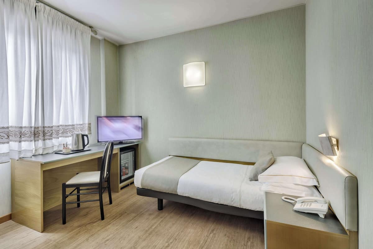 Et hotelværelse med en seng, et skrivebord med computer, en stol, en telefon og et vindue med gardiner.