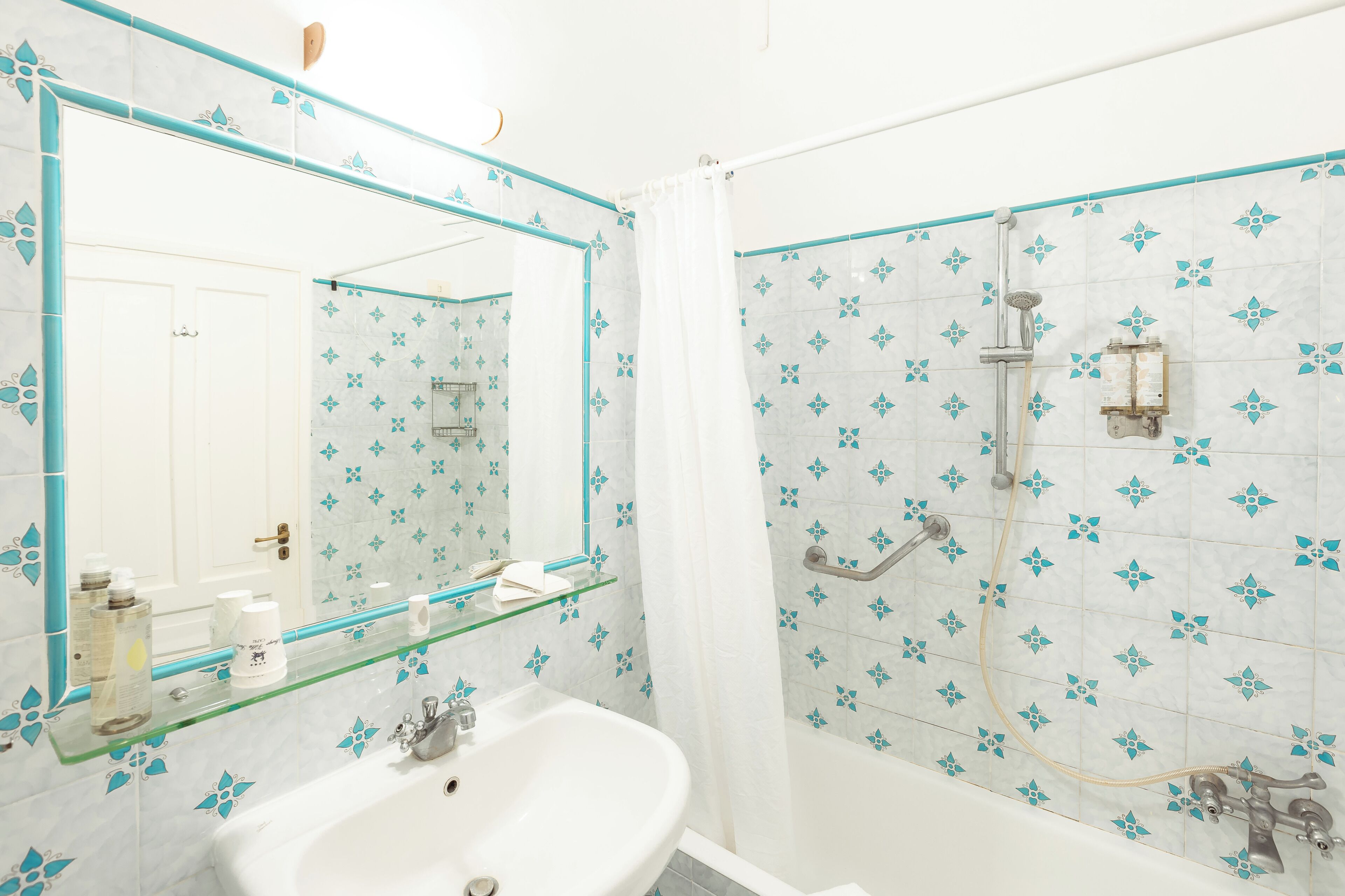 A bathroom with a white sink, a glass shelf, a mirror, and a shower area with blue floral tiles.