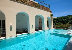 A pool area with clear blue water, a covered seating area with chairs, and a view of a hillside.