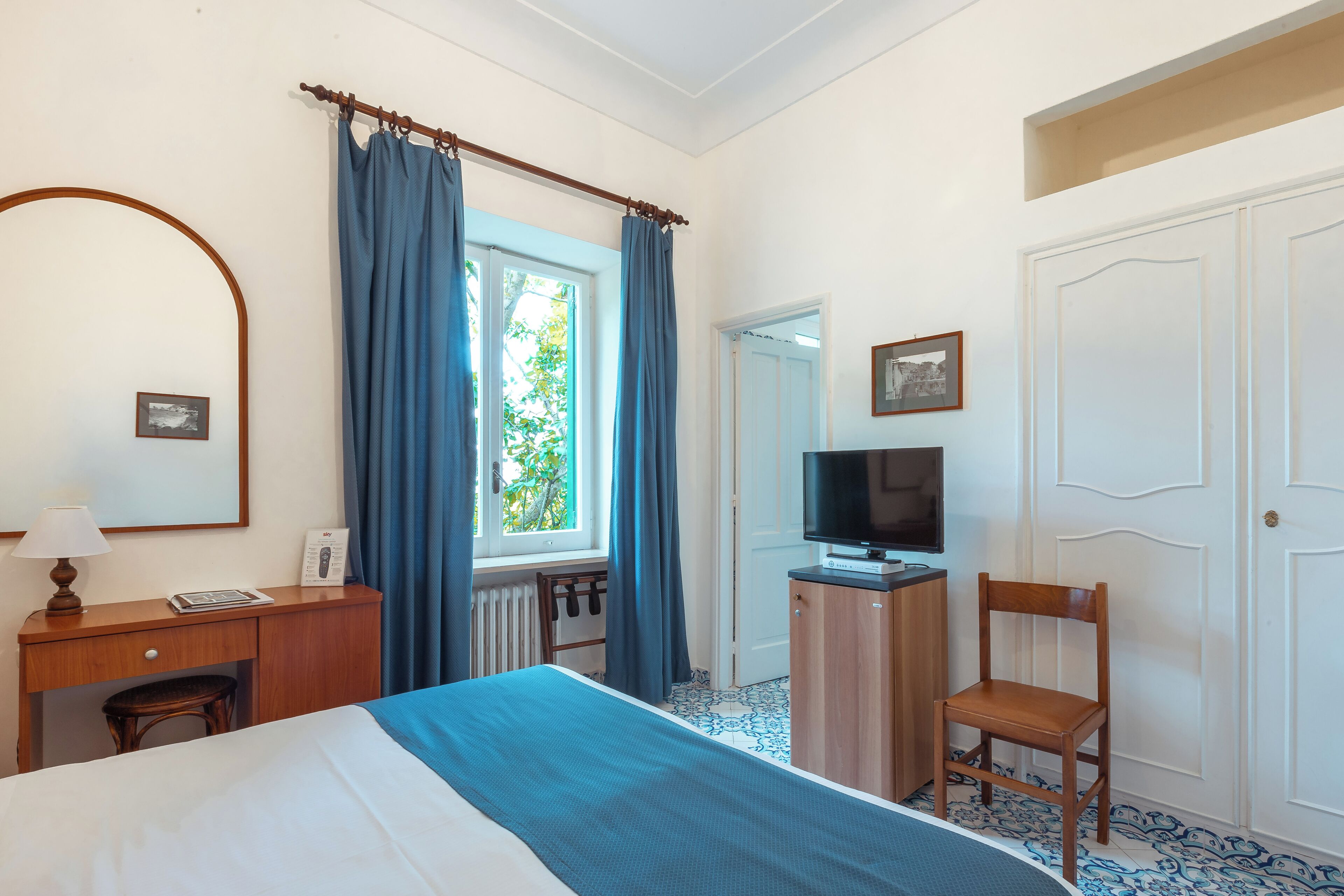Una habitación de hotel con cama, escritorio con lámpara, televisor, silla y ventana con cortinas.