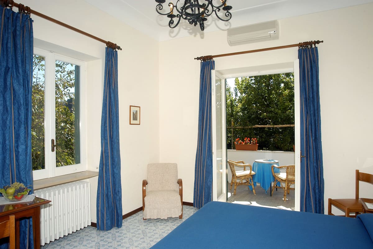 A room with a bed, a chair, a table with a vase, and a view of an outdoor patio with chairs and a table.
