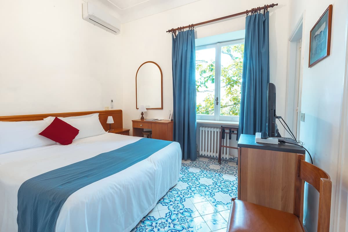 A hotel room with a bed, a desk, a chair, a window with curtains, and a radiator.