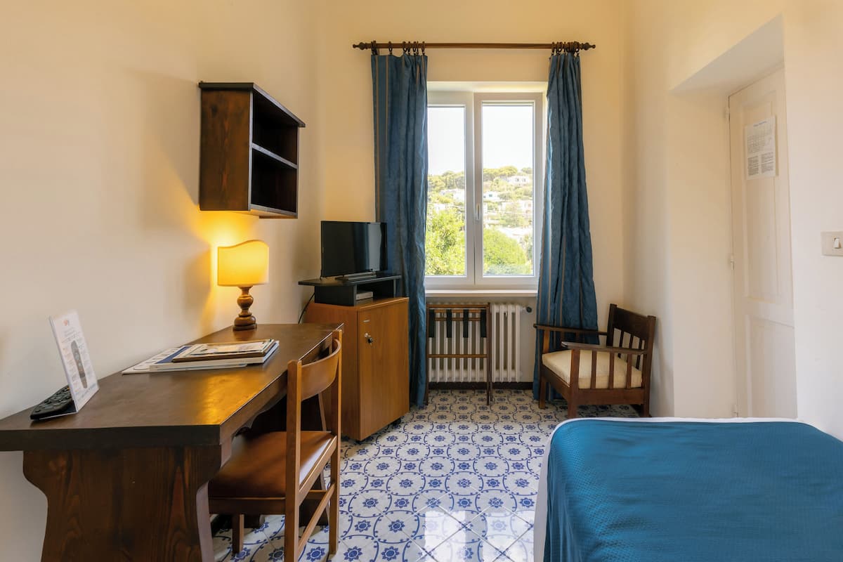 A small room with a wooden desk, a chair, a bed, a radiator, and a window with curtains.