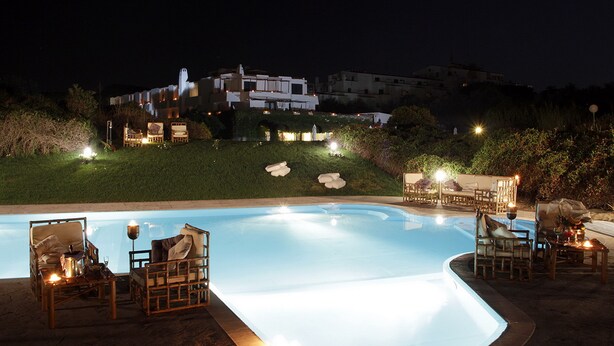 A swimming pool with lounge chairs and tables, surrounded by greenery and a building in the background. - Hotel Della Baia (Cellole)