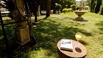 A garden with a statue, a bench, and a book on a table with a glass of orange juice.