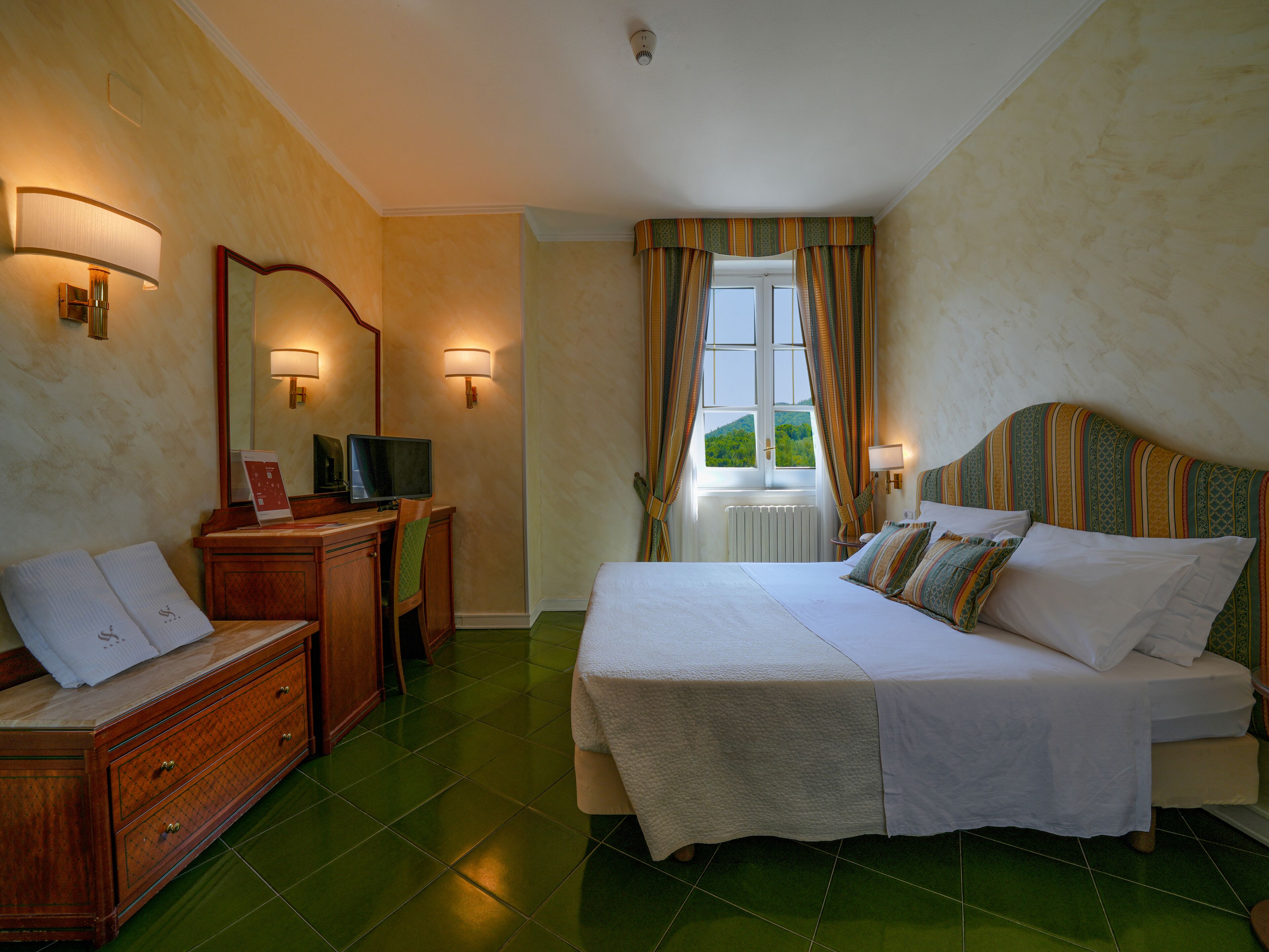 A hotel room with a bed, a desk, a chair, a dresser, and a window with curtains.