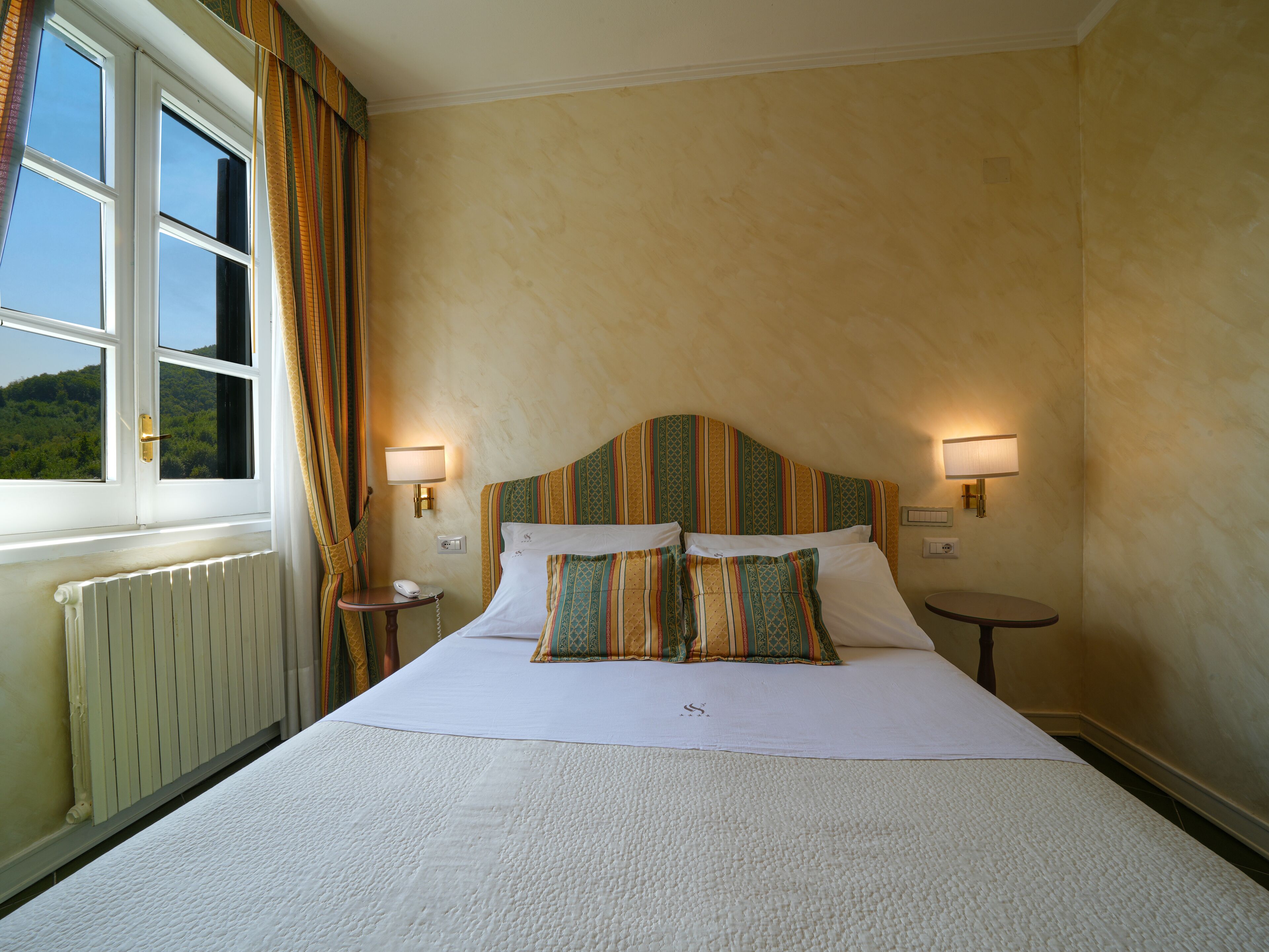 Habitación de hotel con una cama grande, dos mesitas de noche, una ventana con cortinas y vistas al exterior.