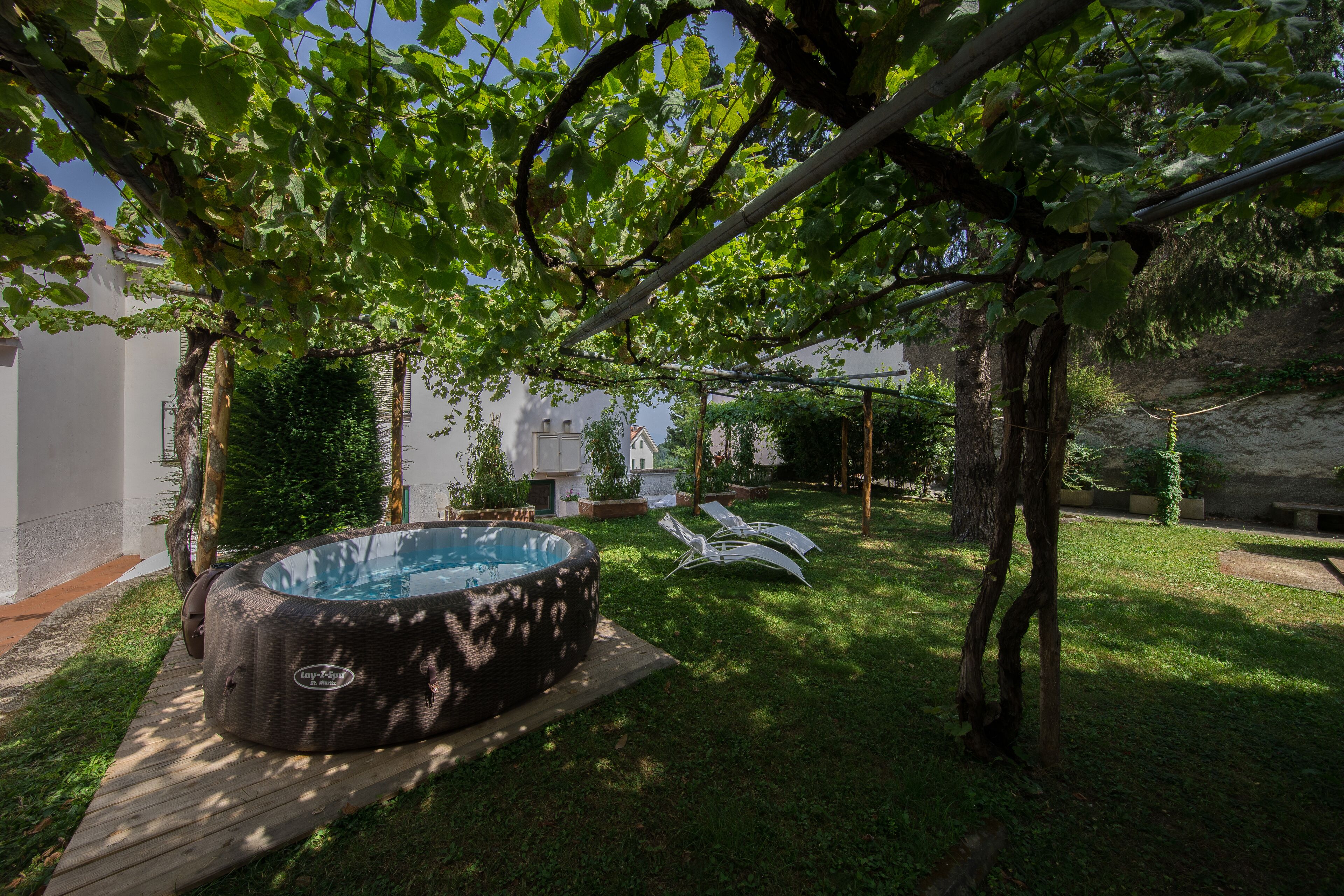 A hot tub under a tree in a backyard with a grassy lawn and a white building in the background.