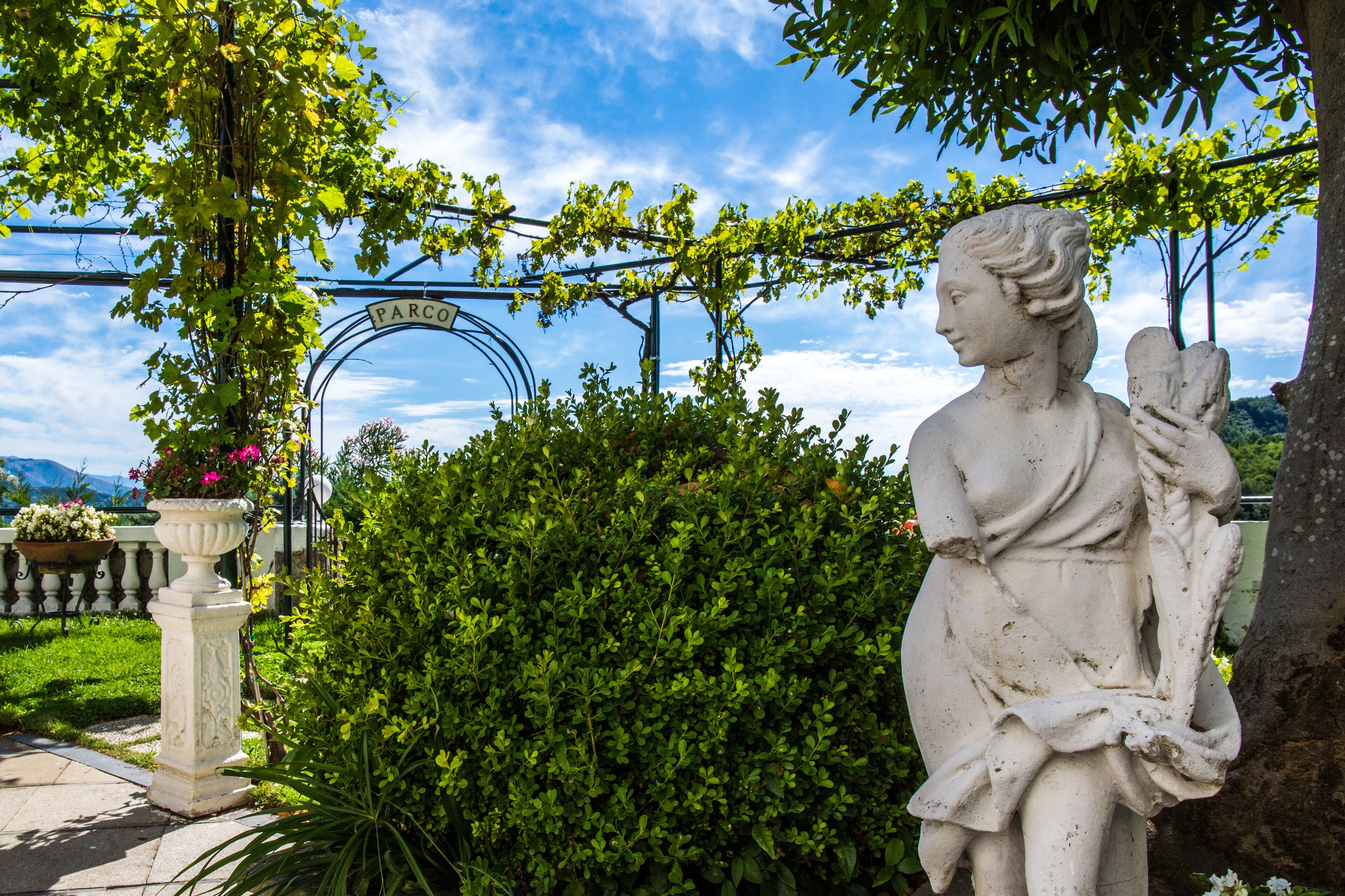 Une statue représentant une femme tenant un enfant, une pergola couverte de plantes grimpantes et un panneau indiquant « PARCO ».