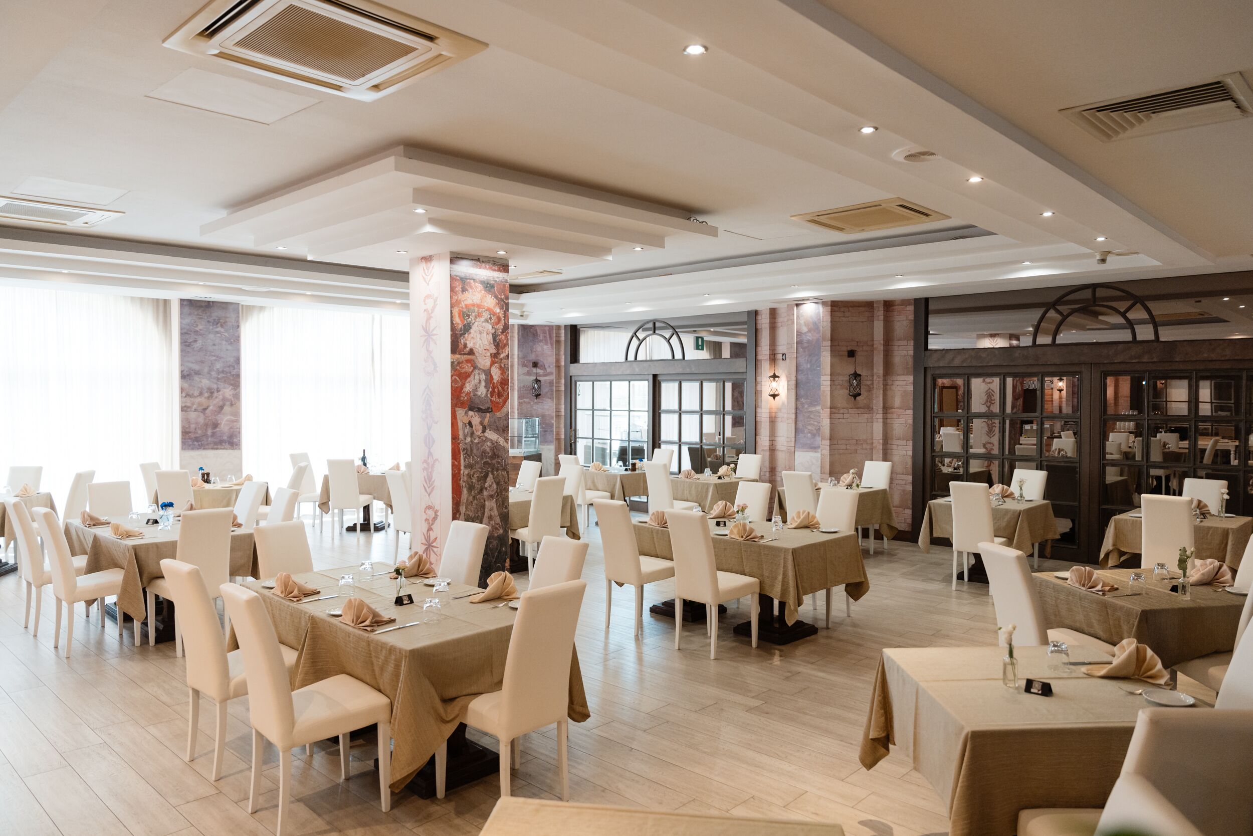 A spacious dining area with multiple tables and chairs, large windows, and a modern ceiling design.