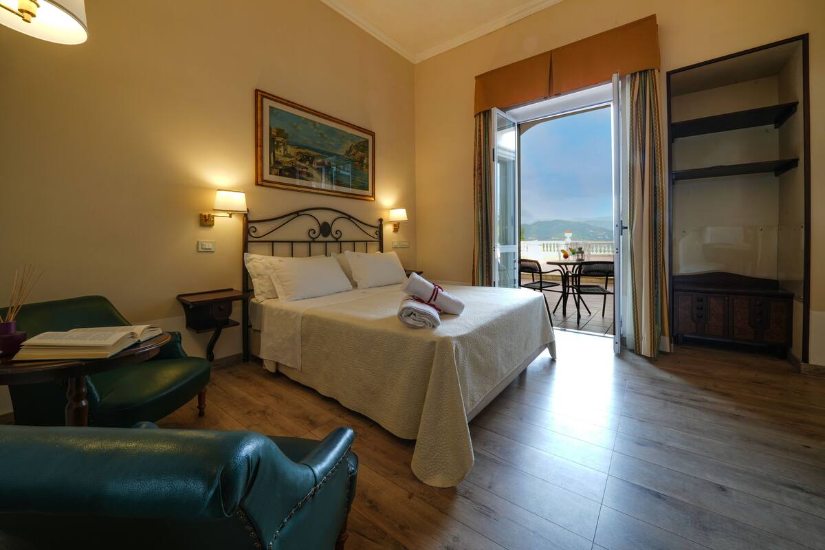 A hotel room with a bed, a desk, a chair, a painting on the wall, and a balcony with a table and chairs.