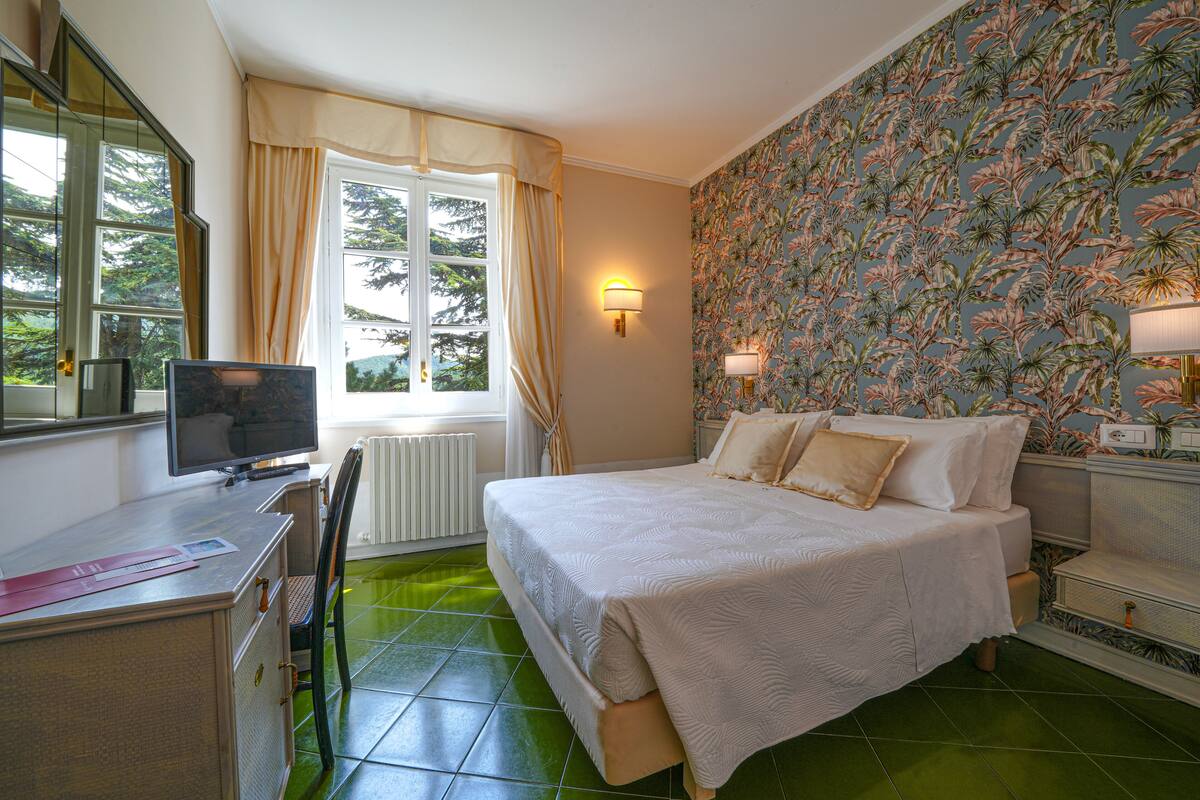 A hotel room with a bed, a desk, a chair, a radiator, a window with curtains, and a wall with a floral wallpaper.