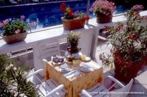 A table set with plates, glasses, and a pineapple on a terrace with potted plants and a pool in the background.