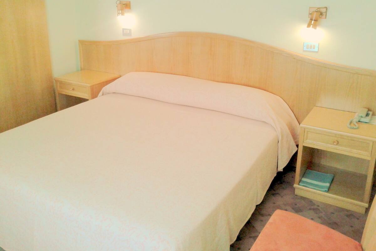Una cama bien tendida con cabecera de madera, dos mesitas de noche con lámparas y un pequeño mueble con cajones y un teléfono.