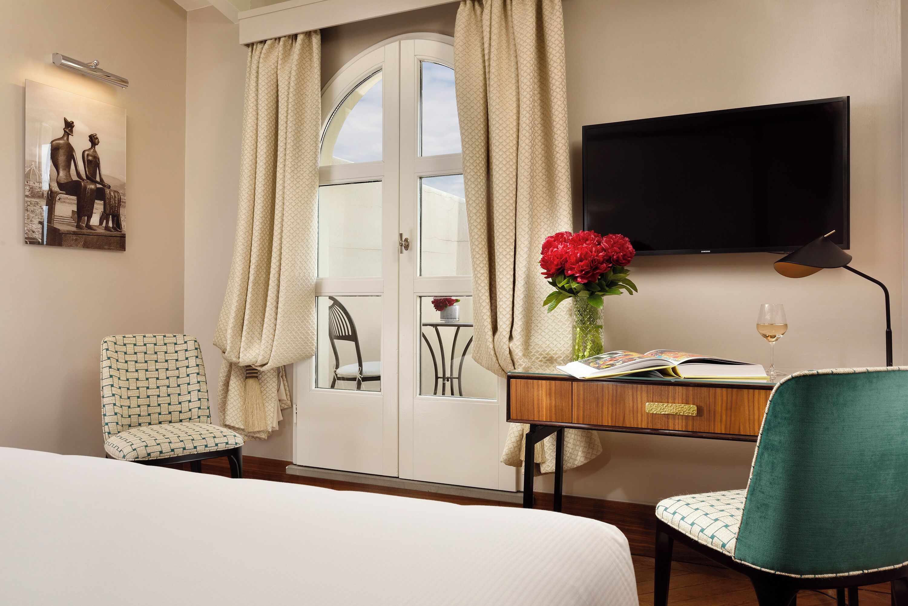 A hotel room with a bed, a desk with a vase of flowers, a chair, a television, and a window with curtains.