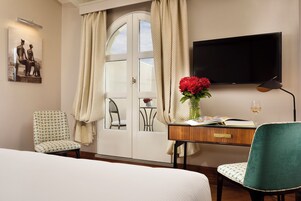 A hotel room with a bed, a desk with a vase of flowers, a chair, a television, and a window with curtains.