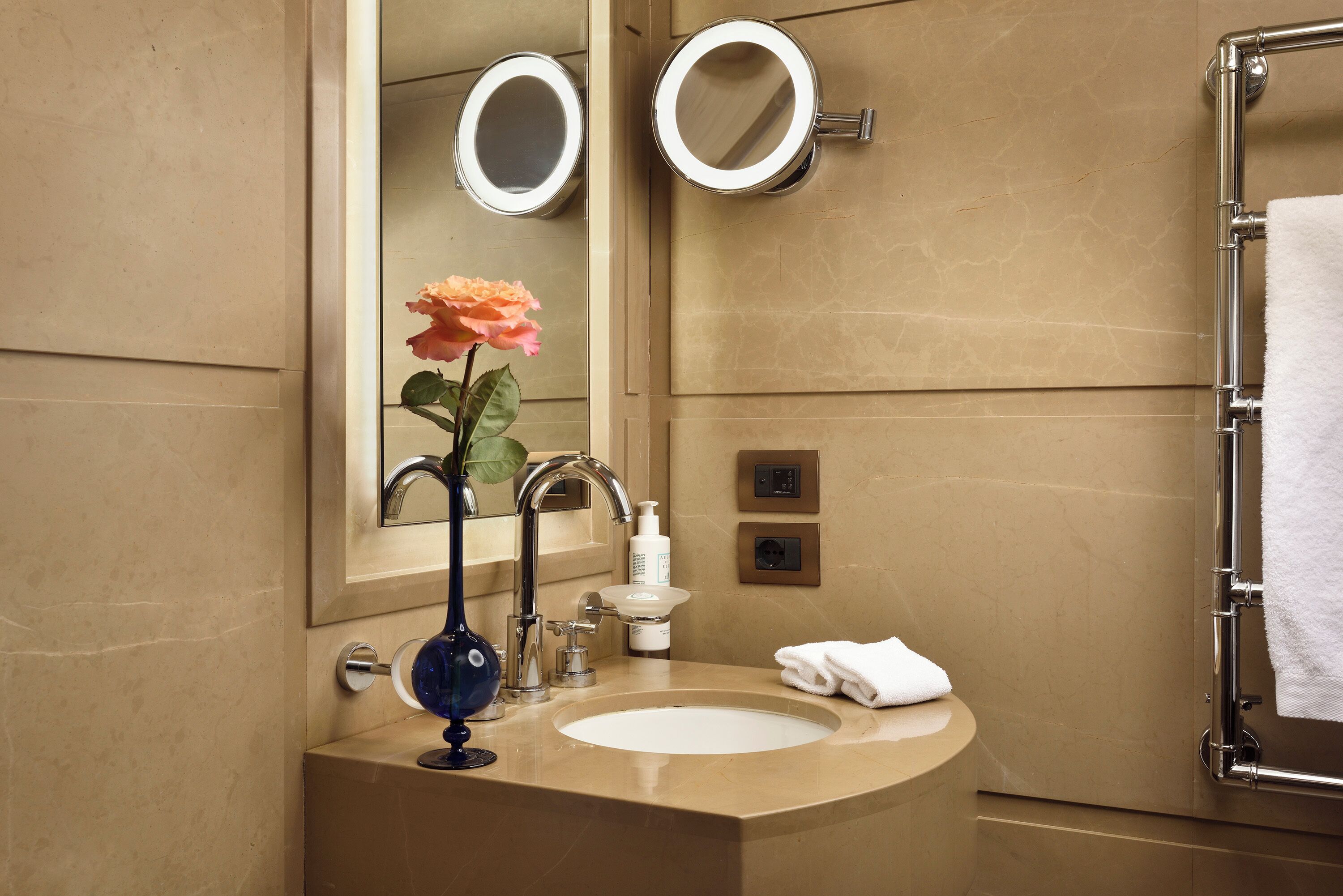 A bathroom with a beige sink, a mirror with two round mirrors, a rose vase, a towel rack, and a soap dispenser.