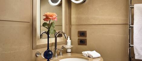 A bathroom with a beige sink, a mirror with two round mirrors, a rose vase, a towel rack, and a soap dispenser.