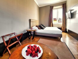 Una camera d'albergo con un letto, un tavolino con una ciotola di frutta, una TV a parete e una finestra con tende.