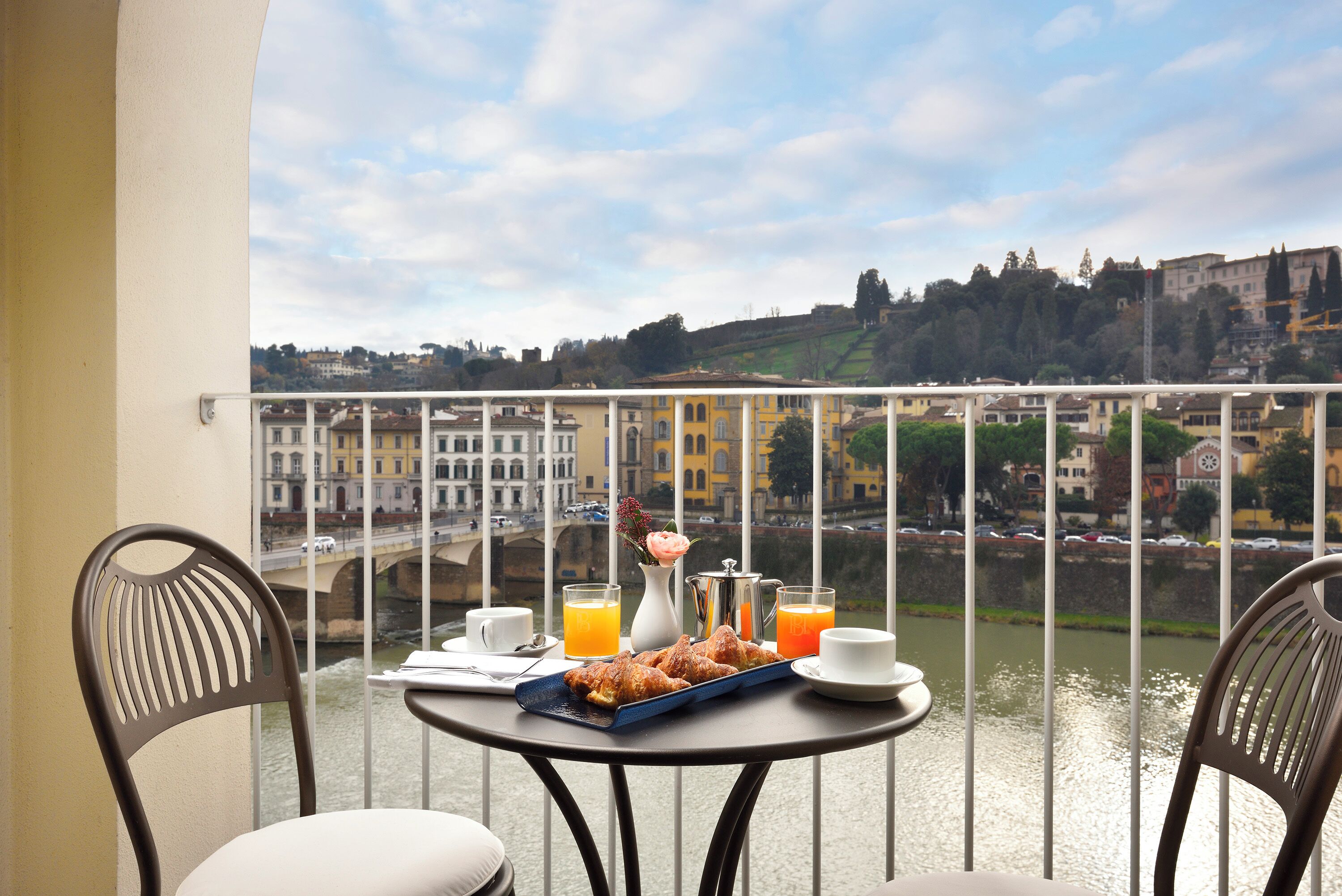 Een balkon met een ontbijttafel, compleet met croissants, sap en koffie, met uitzicht op een rivier en gebouwen.