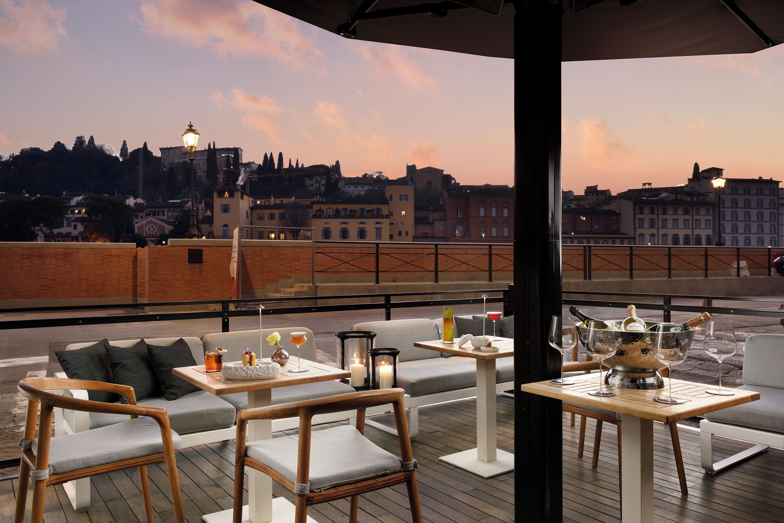 A rooftop terrace with seating, tables, and a view of the cityscape at dusk.