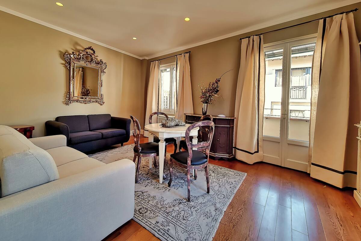 A living room with a sofa, a dining table with chairs, a mirror, and a window with curtains.