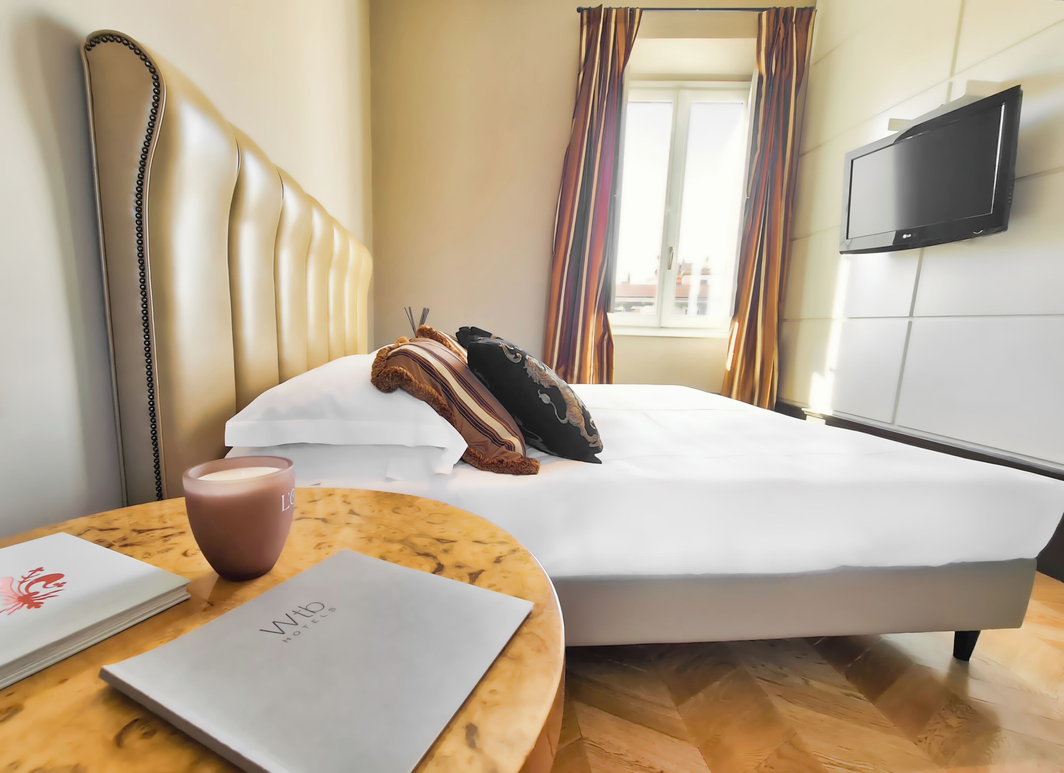 A hotel room with a bed, a bedside table with a cup and a menu, a TV on the wall, and curtains.