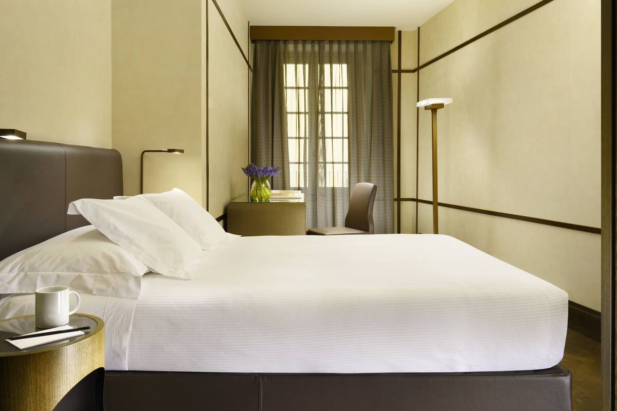 A hotel room with a large bed, a bedside table with a lamp, a chair, and a window with curtains.