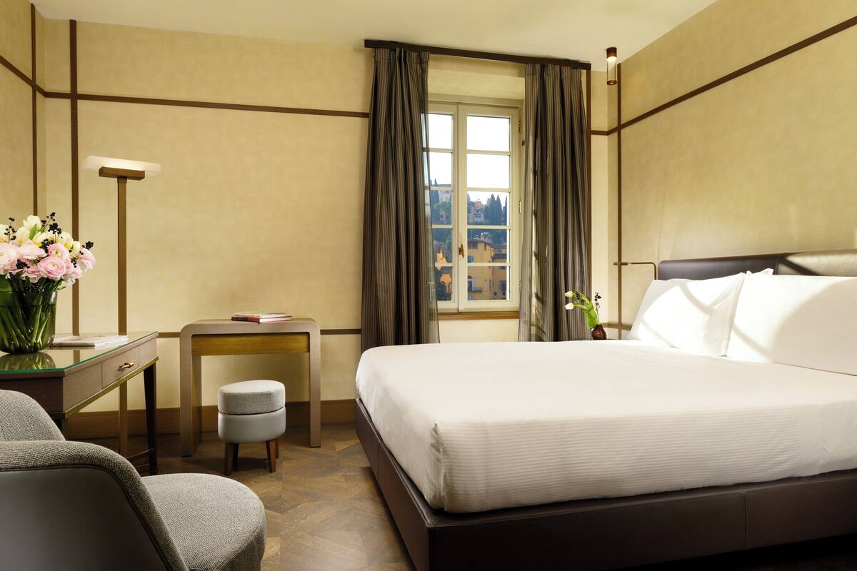 A hotel room with a large bed, a desk, a chair, a small stool, a vase of flowers, and a window with curtains.