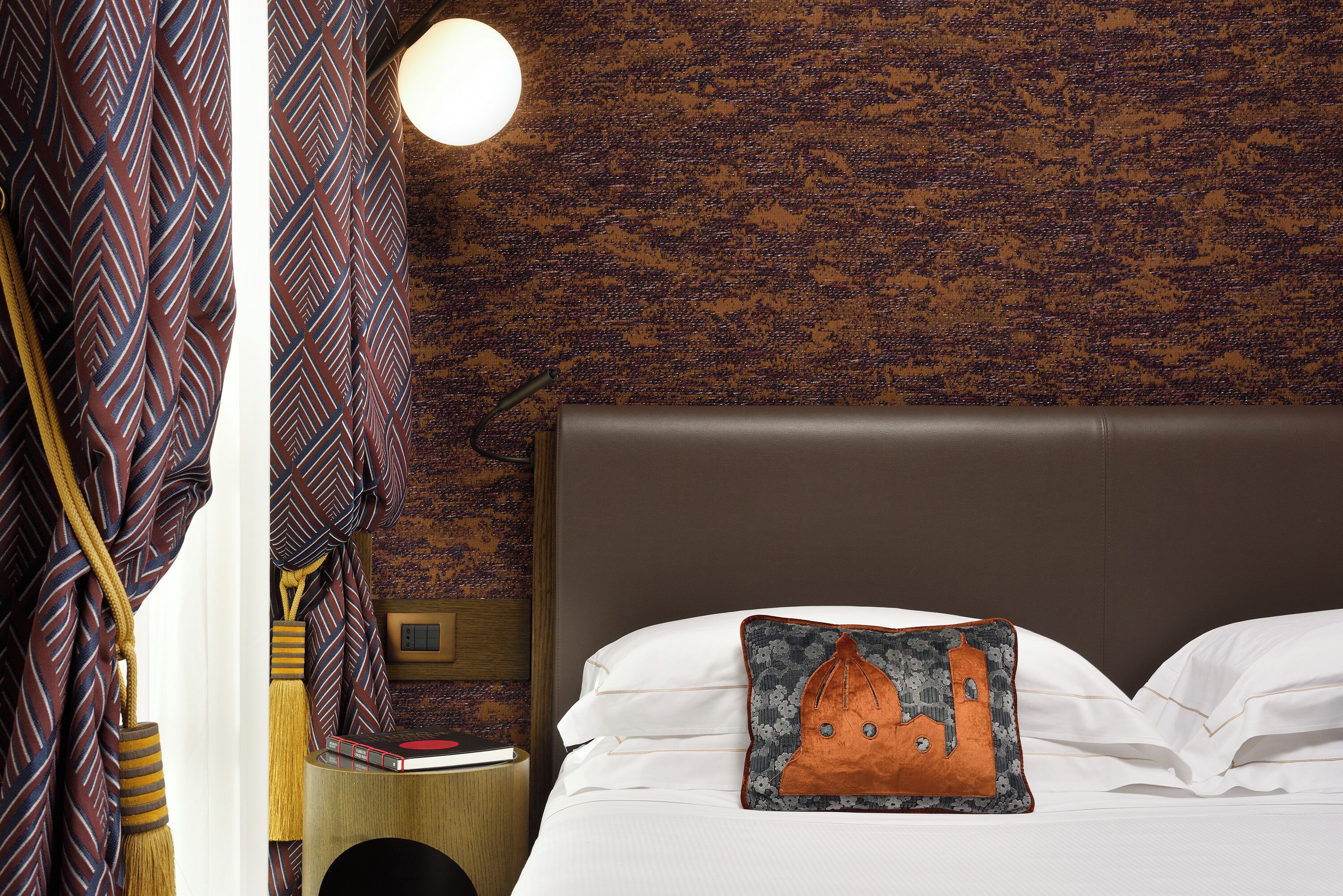 A neatly made bed with a pillow featuring a building design, a bedside lamp, and a wall with a textured pattern.