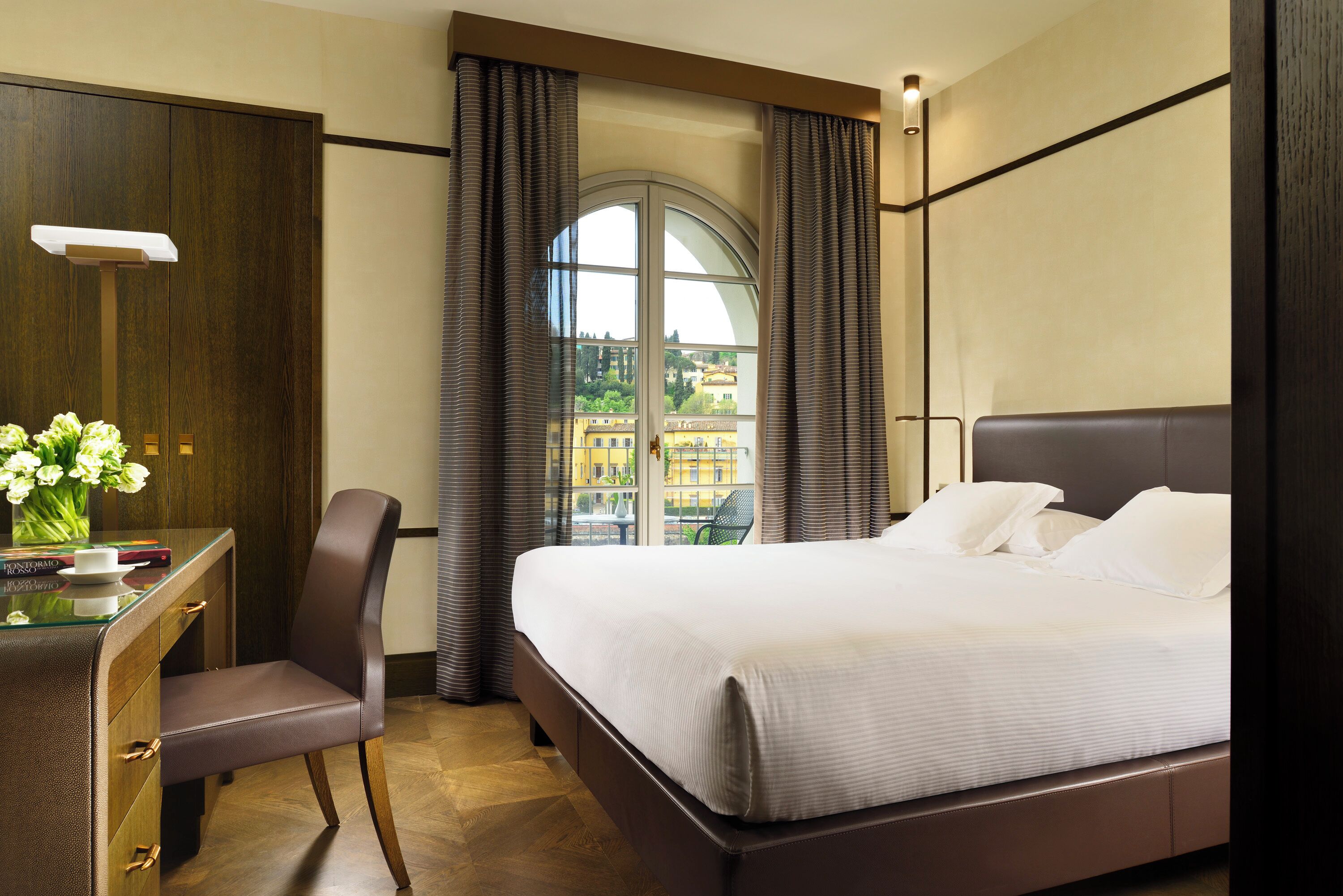 A hotel room with a large bed, a desk with a chair, and a view of buildings through a window with curtains.