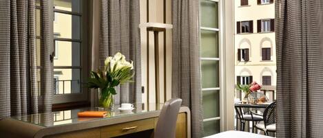 A hotel room with a large window, a desk with a vase of flowers, a chair, and a view of buildings outside.