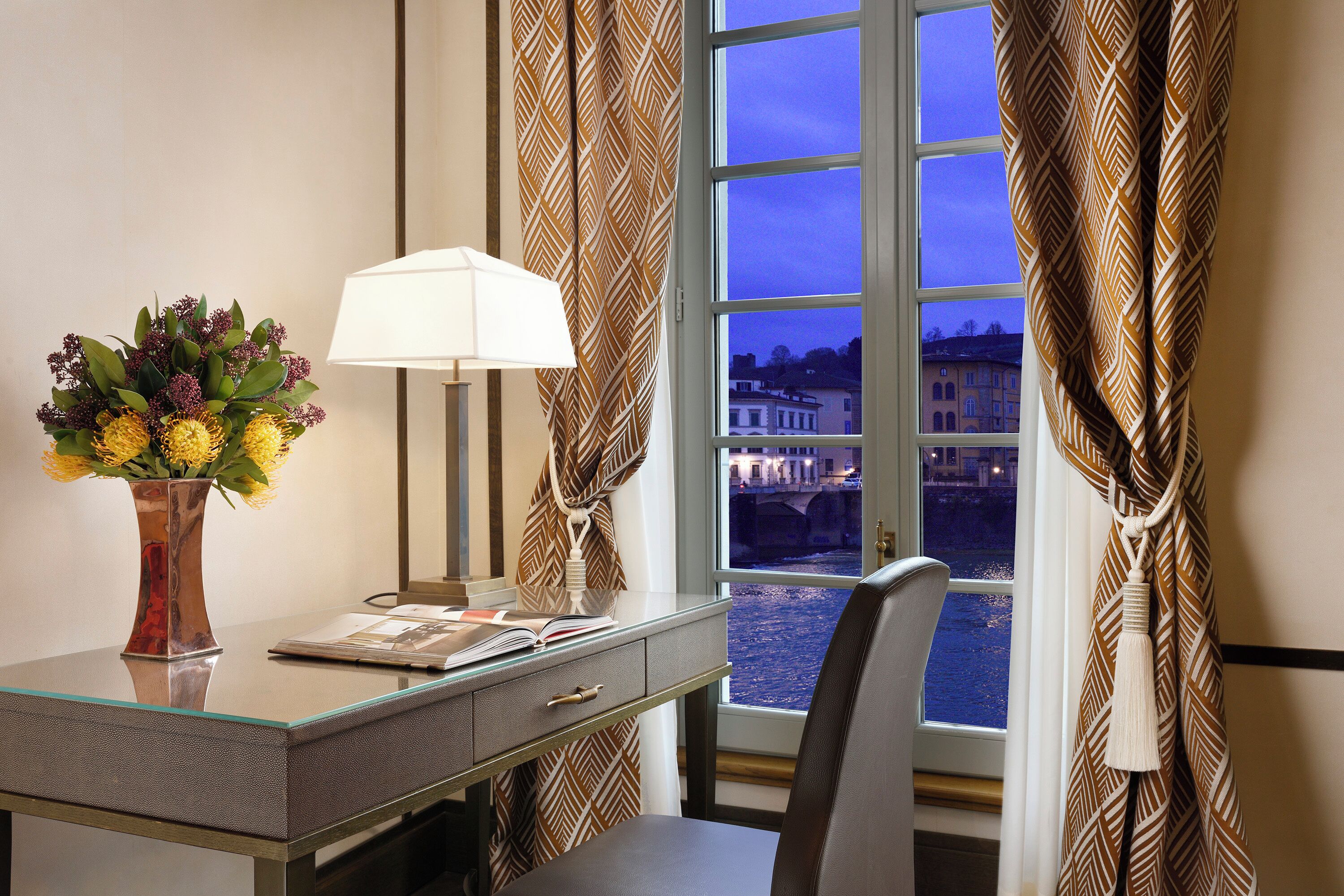 A hotel room with a desk, a lamp, a vase with flowers, and a view of a bridge through the window.