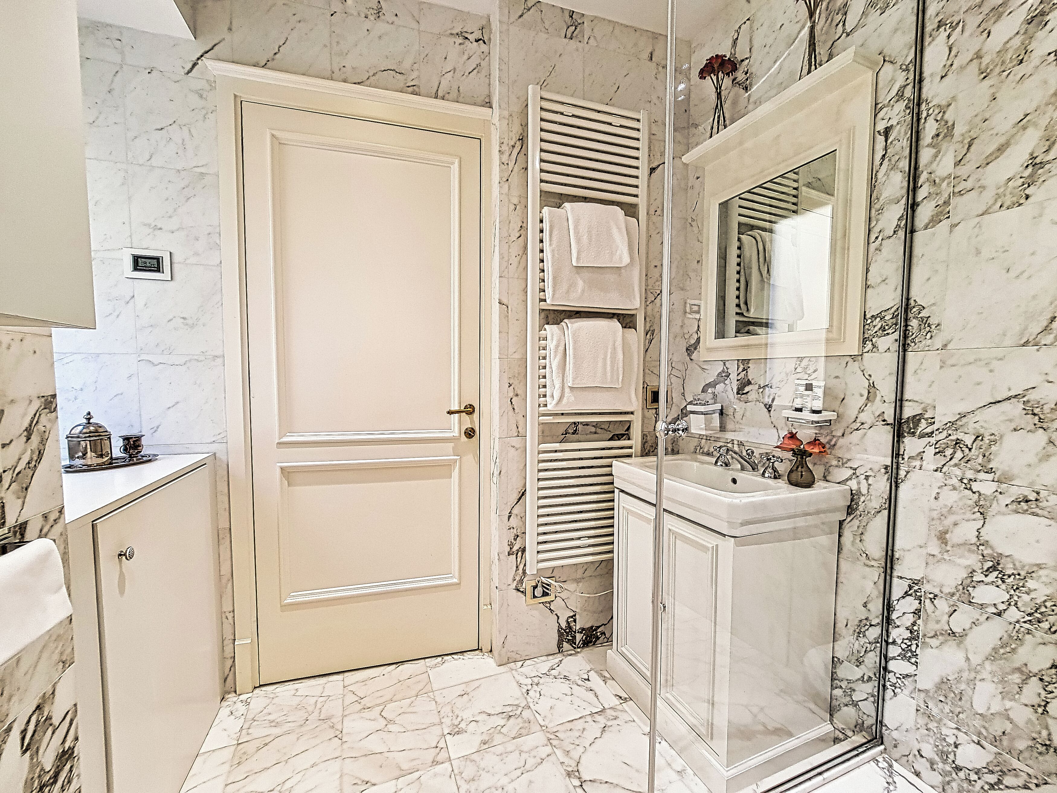 A bathroom with a marble floor, a glass shower enclosure, a sink with toiletries, and a towel rack.