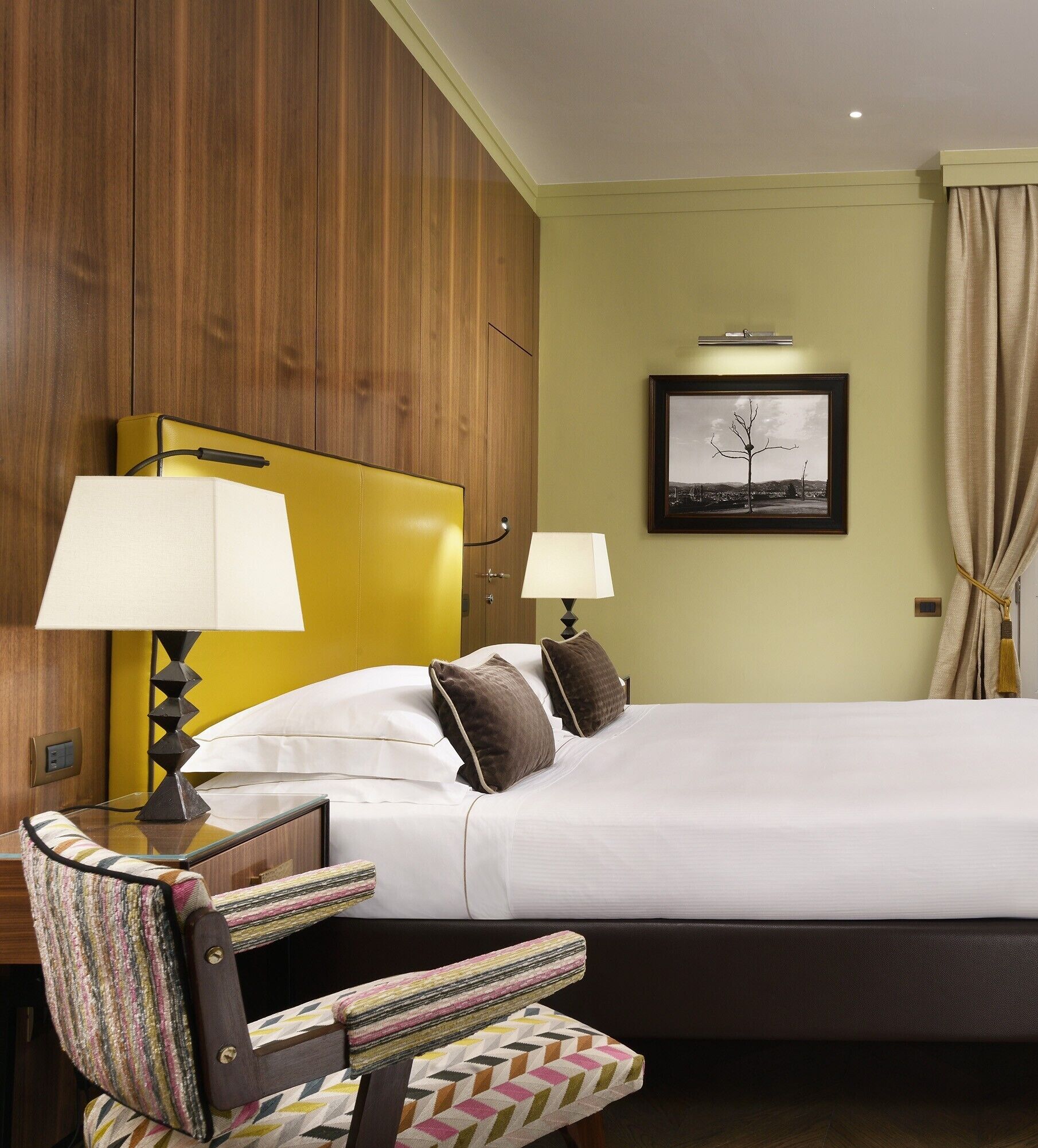 A hotel room with a bed, two bedside lamps, a framed picture on the wall, and a chair with a colorful cushion.