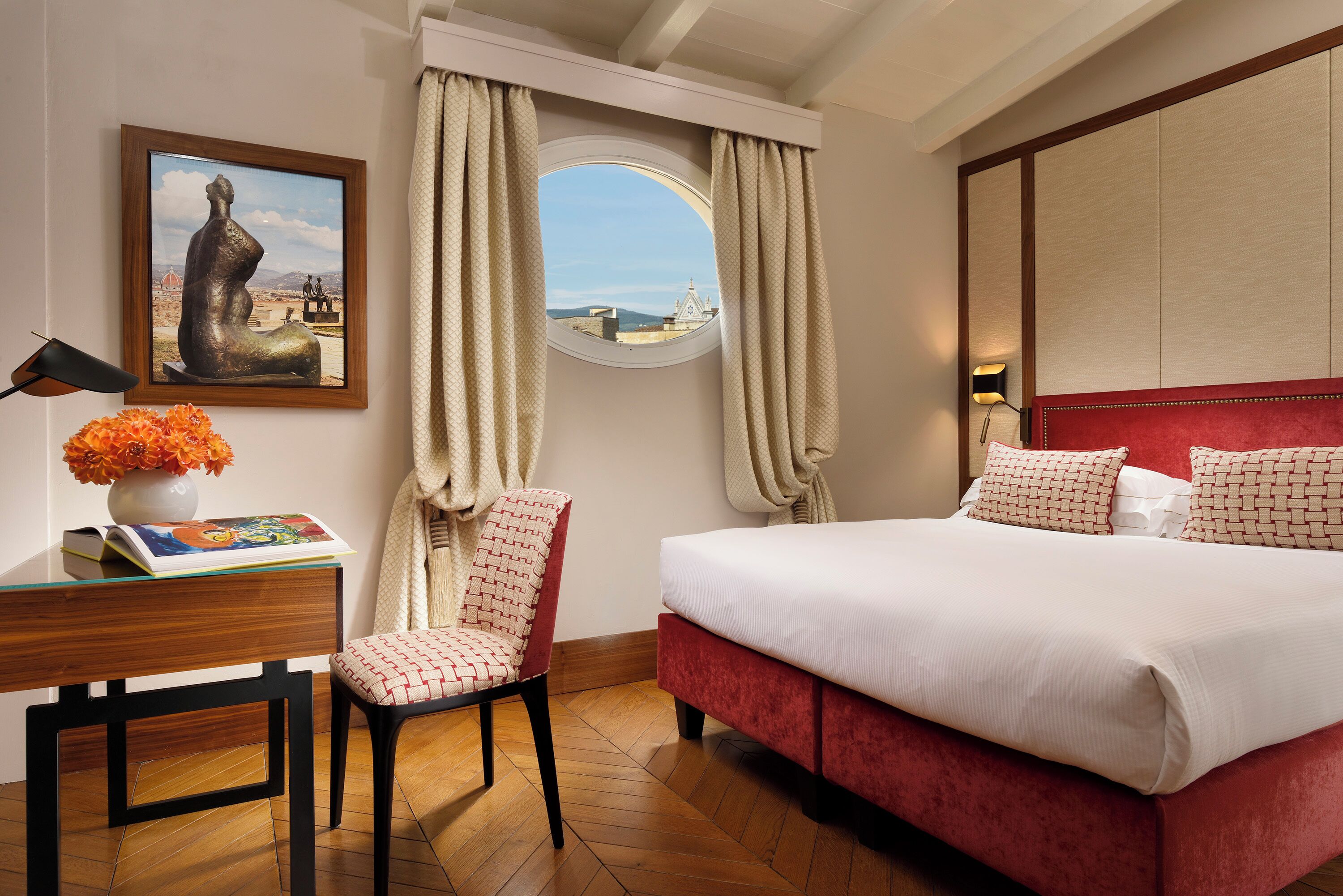 A hotel room with a bed, a desk with a lamp, a chair, a window with curtains, and a painting on the wall.