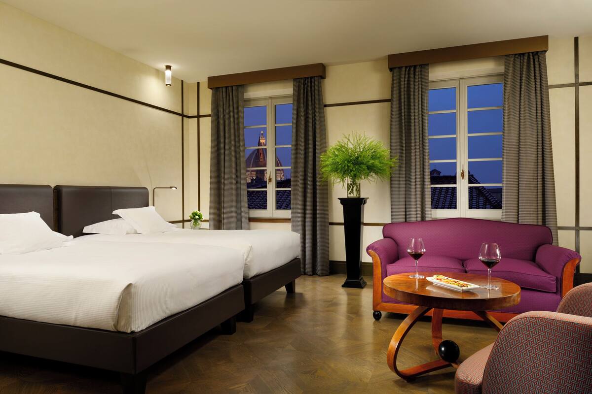 A hotel room with a large bed, a sofa, a small table with glasses of wine, and a potted plant.