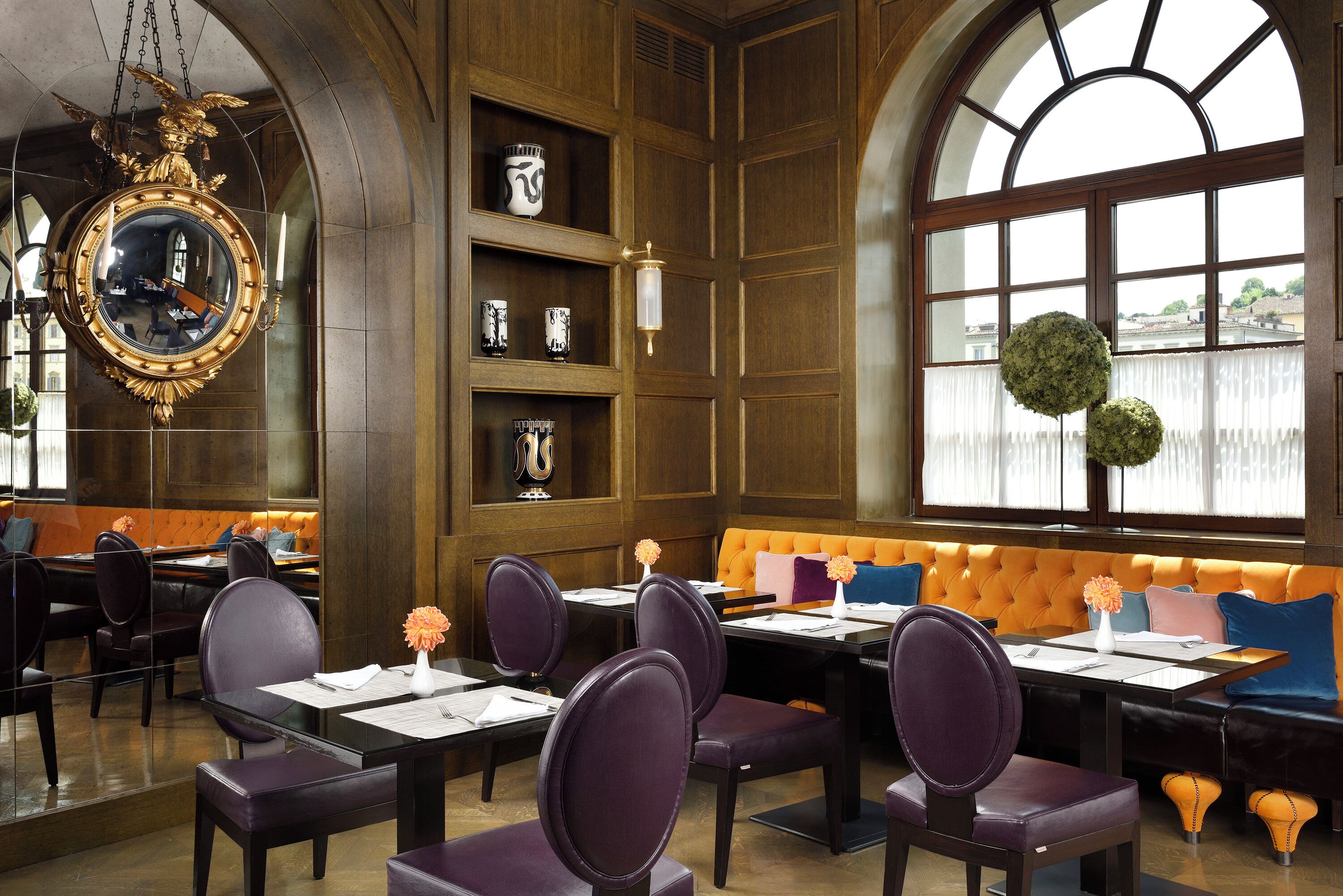 A restaurant with a high arched window, a large mirror, and a seating area with purple chairs and an orange cushioned bench.