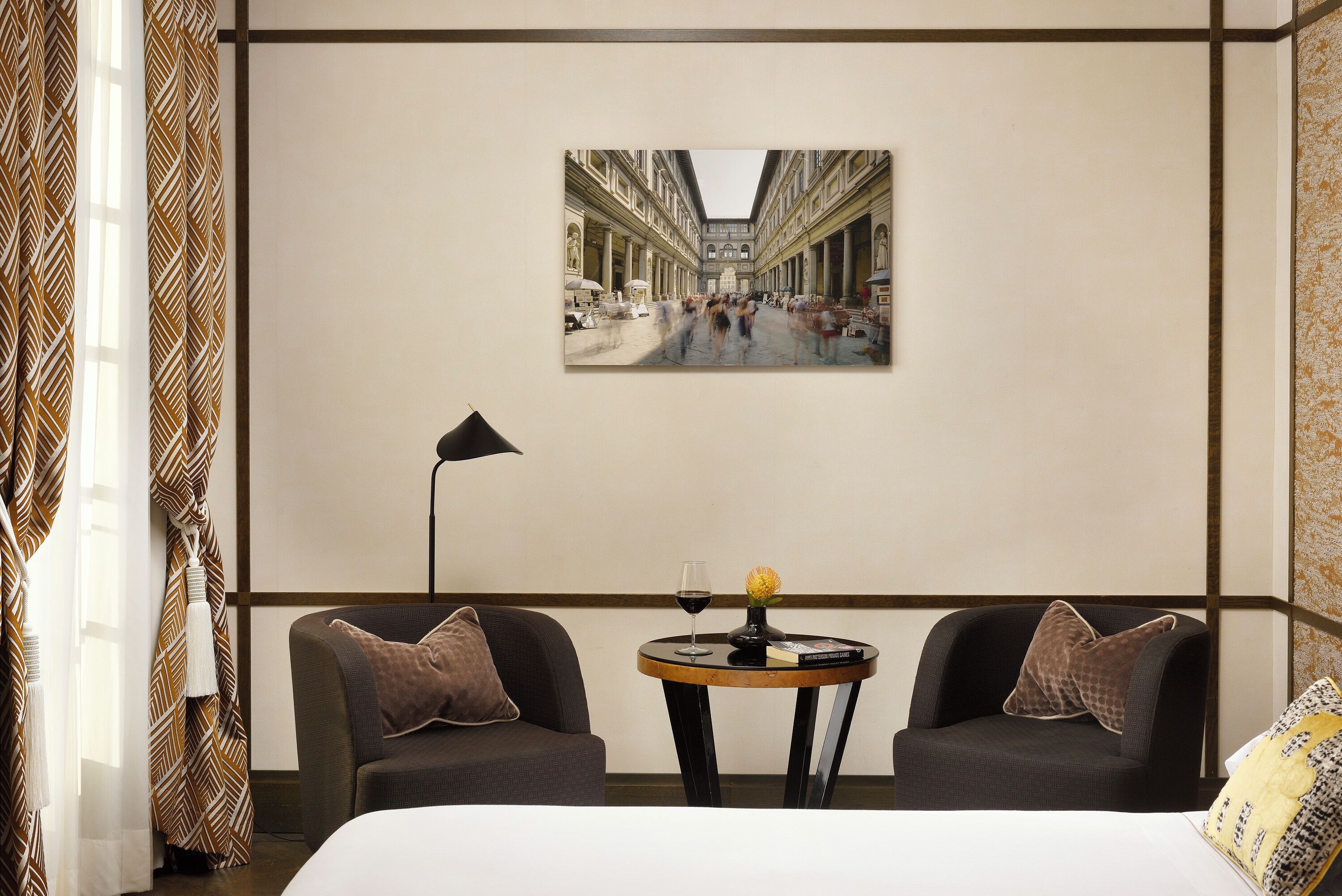 Une chambre d’hôtel avec un lit, deux fauteuils, une petite table ronde avec un verre à vin et un vase, et un grand tableau représentant une scène de rue accroché au mur.