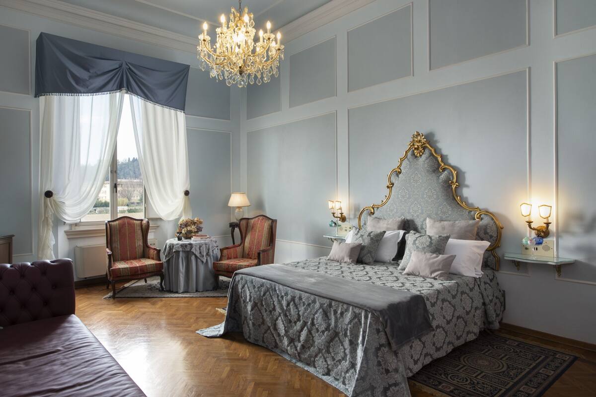 A bedroom with a large bed, a chandelier, a window with curtains, a chair, and a nightstand.